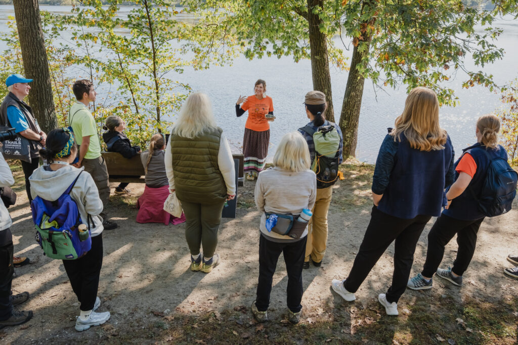A person leads an outdoor workshop in front of a tree-lined river.